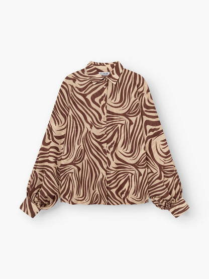 Die Antonia Voile Zebra Brown ist eine langärmelige Bluse mit braunem und beigem Zebramuster, Kragen und gerafften Manschetten. Die in Italien gefertigte Bluse besticht durch ihren Stil und ihre hochwertige Verarbeitung und wird flach auf weißem Grund gezeigt.