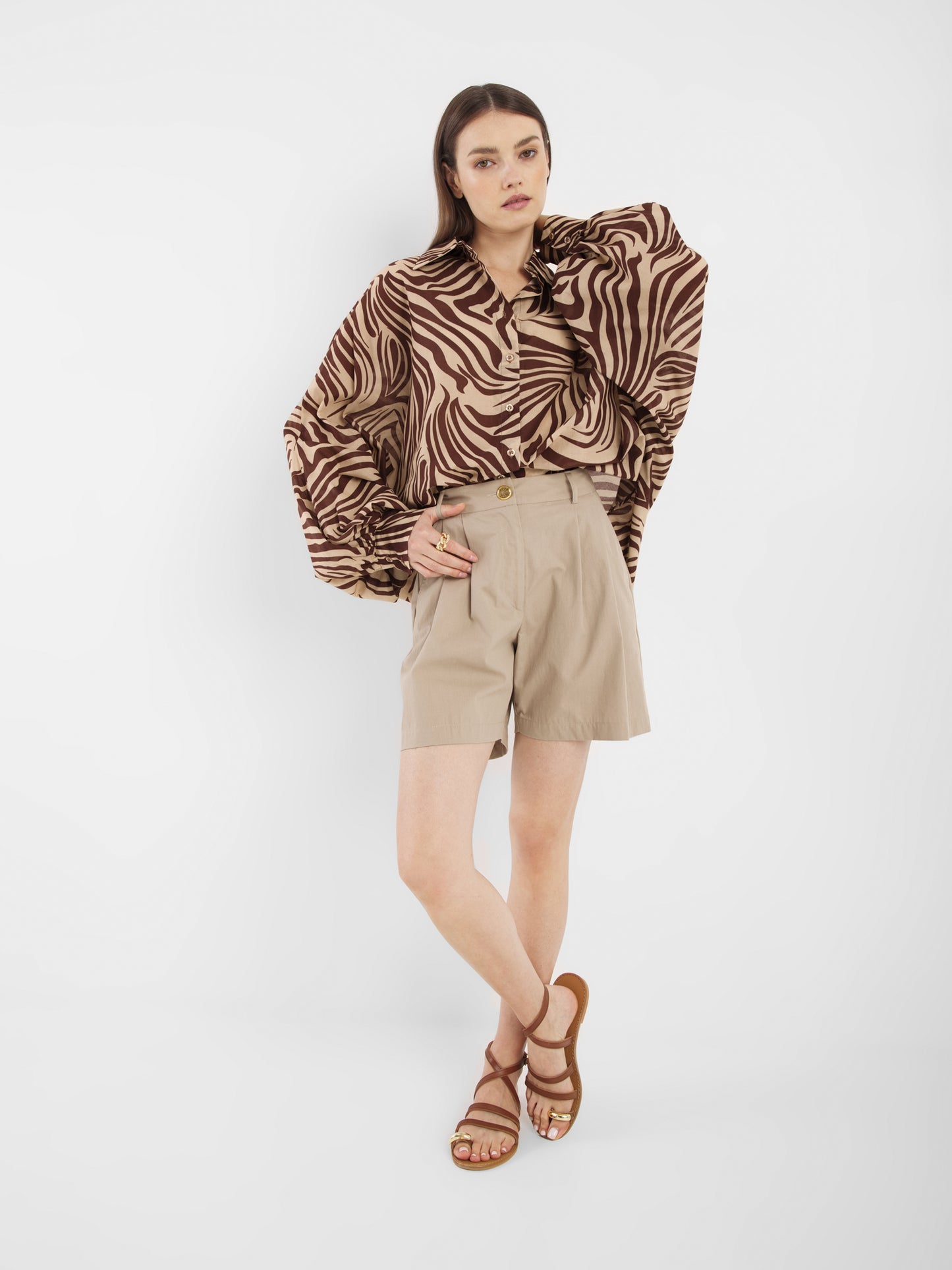 Eine Frau posiert vor einem weißen Hintergrund und trägt die Antonia Voile Zebra Brown Bluse mit übergroßen Ärmeln, beigefarbene Shorts mit hoher Taille und braune Riemchensandalen - ein mühelos schicker Look, Made in Italy.