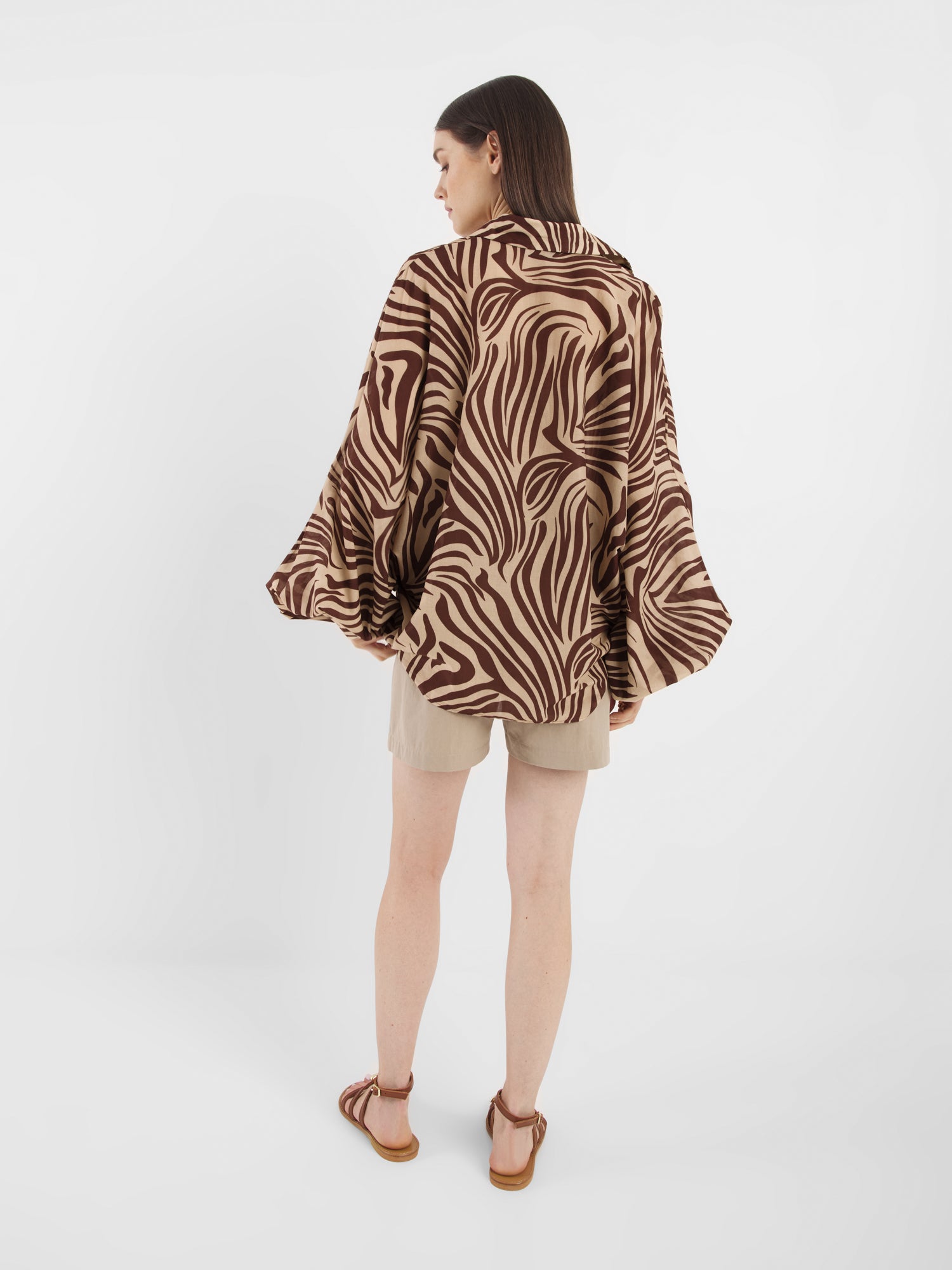 Eine Frau schaut weg und trägt die Antonia Voile Zebra Brown Bluse mit hellbraunen Shorts und braunen Sandalen vor einem schlichten weißen Hintergrund. Diese mühelos schicke Statement-Bluse wird in Italien hergestellt.