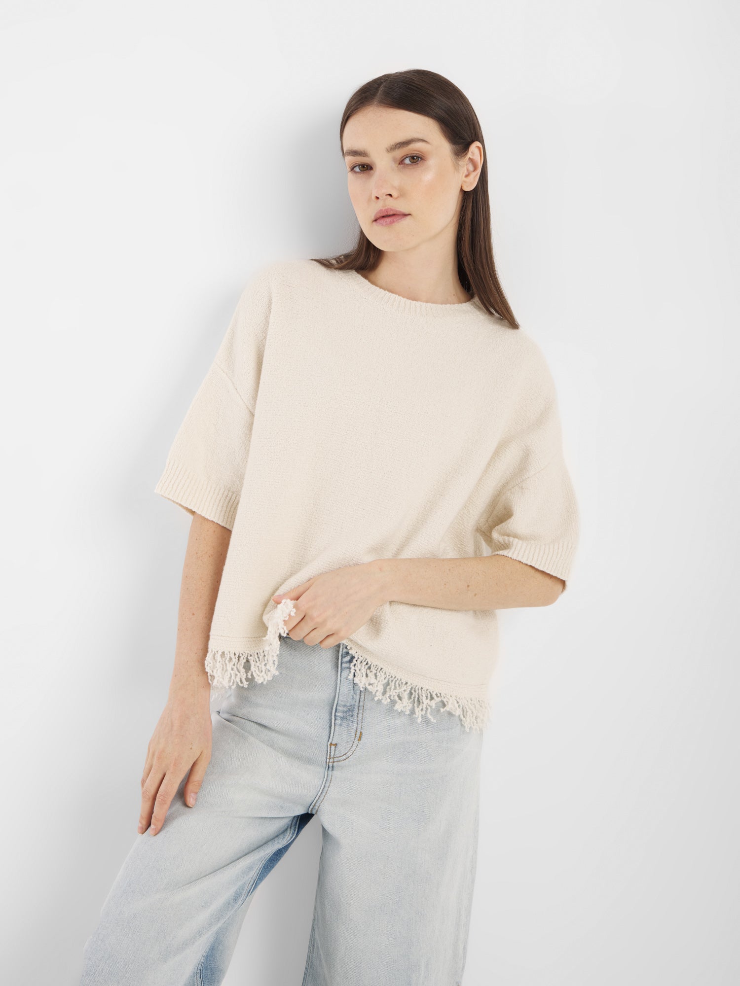 Eine Frau mit glattem braunem Haar, bekleidet mit dem Aurora Fringe Panna - einem locker sitzenden cremefarbenen Strick-Sommer-Top mit Spitzensaum aus Bio-Baumwoll-Mix Damen-Stoff - und hellblauen Jeans steht vor einem schlichten weißen Hintergrund.