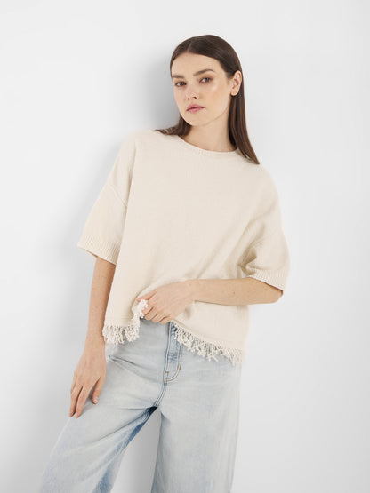 Eine Frau mit glattem braunem Haar, bekleidet mit dem Aurora Fringe Panna - einem locker sitzenden cremefarbenen Strick-Sommer-Top mit Spitzensaum aus Bio-Baumwoll-Mix Damen-Stoff - und hellblauen Jeans steht vor einem schlichten weißen Hintergrund.