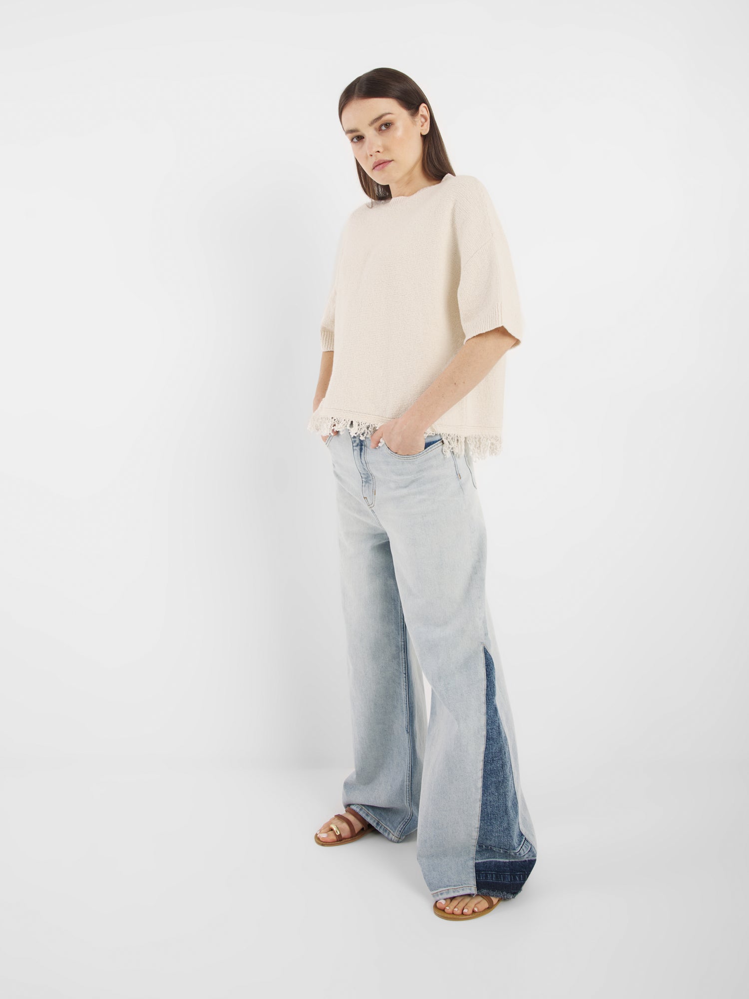 Eine Frau trägt das Aurora Fringe Panna lockeres Strickshirt aus Bio-Baumwoll-Mix mit Fransen am Saum, kombiniert mit hellblauen weiten Jeans und braunen Sandalen. Sie steht vor weißem Hintergrund, die Hände in den Taschen.