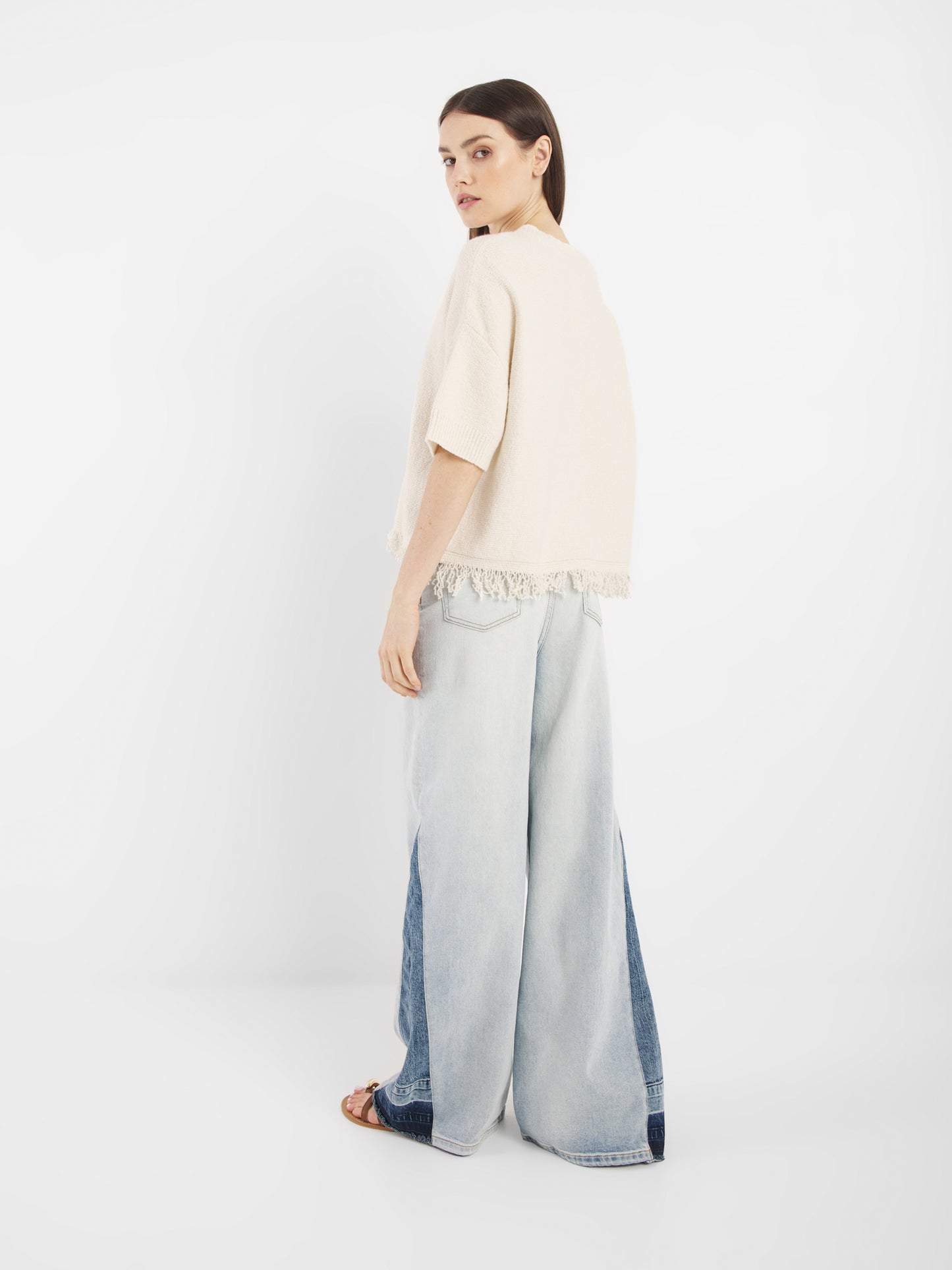 Eine Frau steht vor einem weißen Hintergrund, schaut nach hinten und wirft einen Blick über ihre Schulter. Sie trägt das Oberteil Aurora Fringe Panna mit Fransen und sehr weit geschnittene, zweifarbige hellblaue Jeans mit Sandalettenabsätzen.