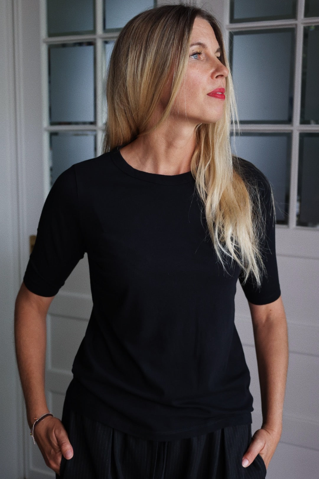 Eine Frau mit langen blonden Haaren trägt das Half Sleeve Black Shirt, steht in einem Haus und schaut nach rechts. Im Hintergrund sind ein Fenster mit Milchglasscheiben und eine weiße Tür zu sehen.