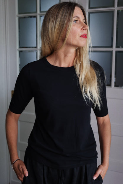 Eine Frau mit langen blonden Haaren trägt das Half Sleeve Black Shirt, steht in einem Haus und schaut nach rechts. Im Hintergrund sind ein Fenster mit Milchglasscheiben und eine weiße Tür zu sehen.