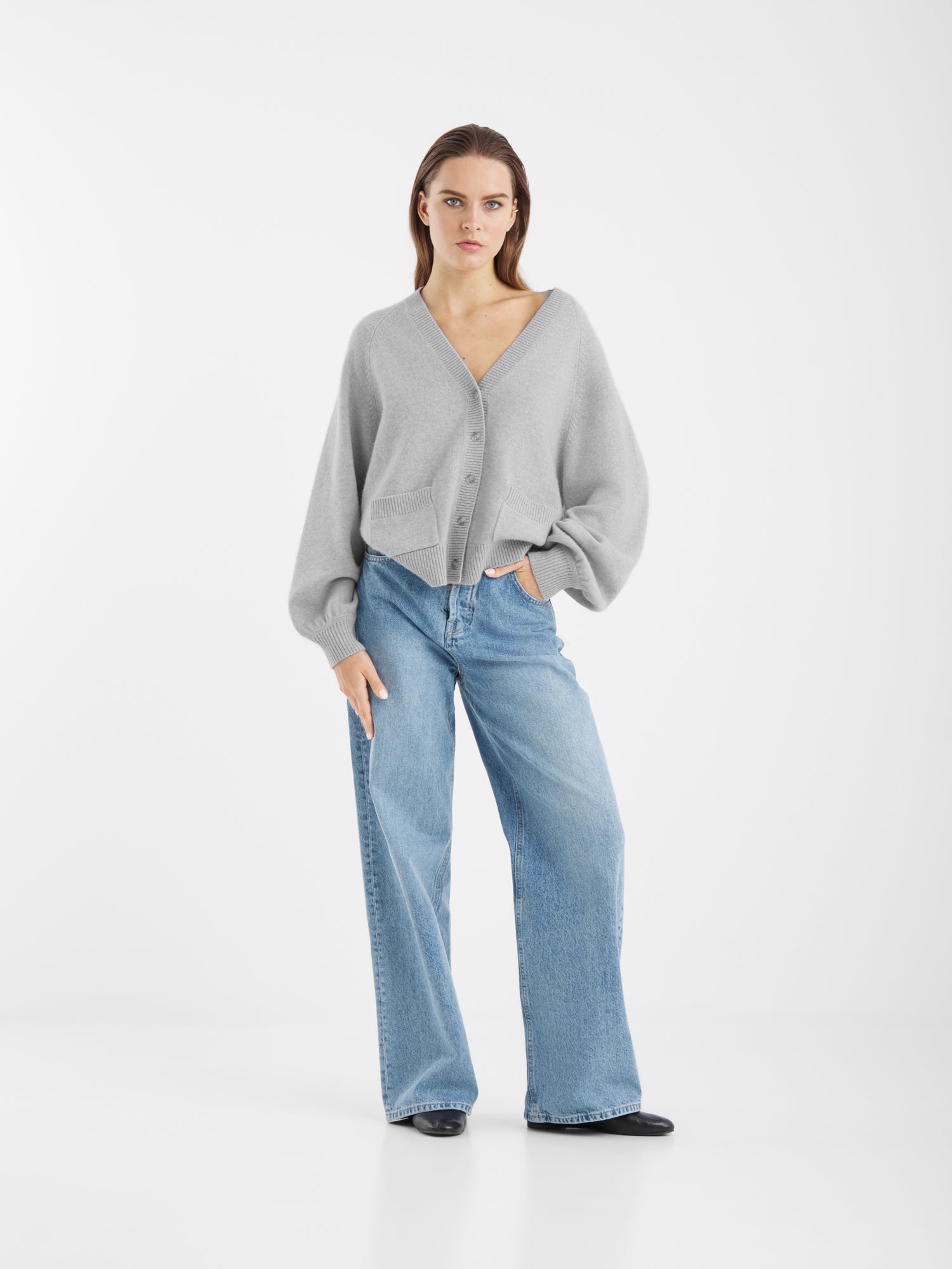 Ein weibliches Modell steht vor einem weißen Hintergrund und trägt den Bella Cardigan Light Grey, hellblaue Jeans mit weitem Bein und schwarze Schuhe. Sie hat die linke Hand in der Tasche und einen neutralen Gesichtsausdruck.