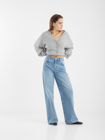 Eine Frau steht neutral vor weißem Hintergrund und trägt den Bella Cardigan Light Grey sowie eine hoch taillierte, weit geschnittene blaue Jeans und schwarze Schuhe.