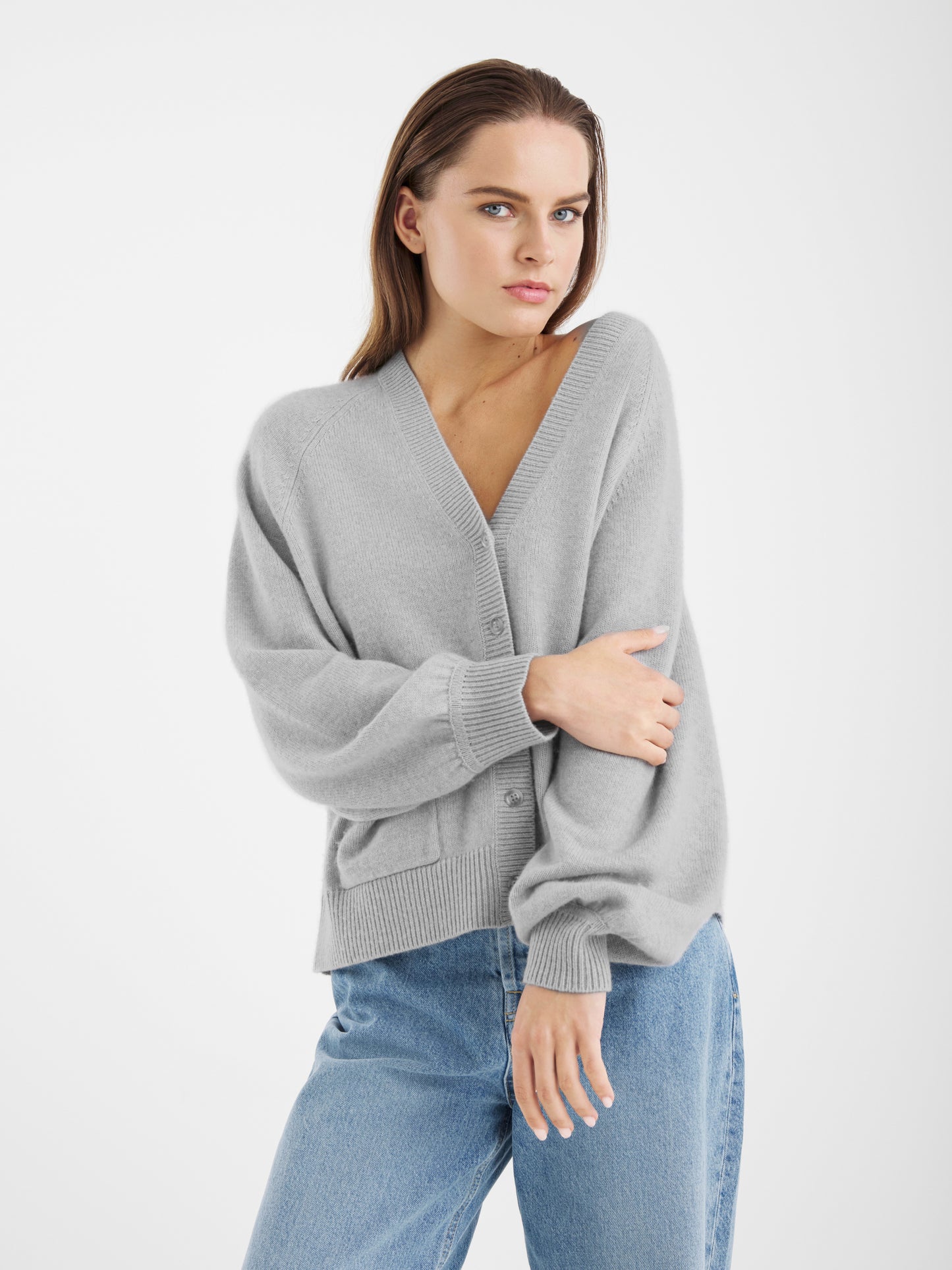Ein Damen Modell trägt den Bella Cardigan Light Grey und blaue Jeans, steht vor weißem Hintergrund, blickt in die Kamera, hat einen Arm verschränkt und lässt den anderen an ihrer Seite.