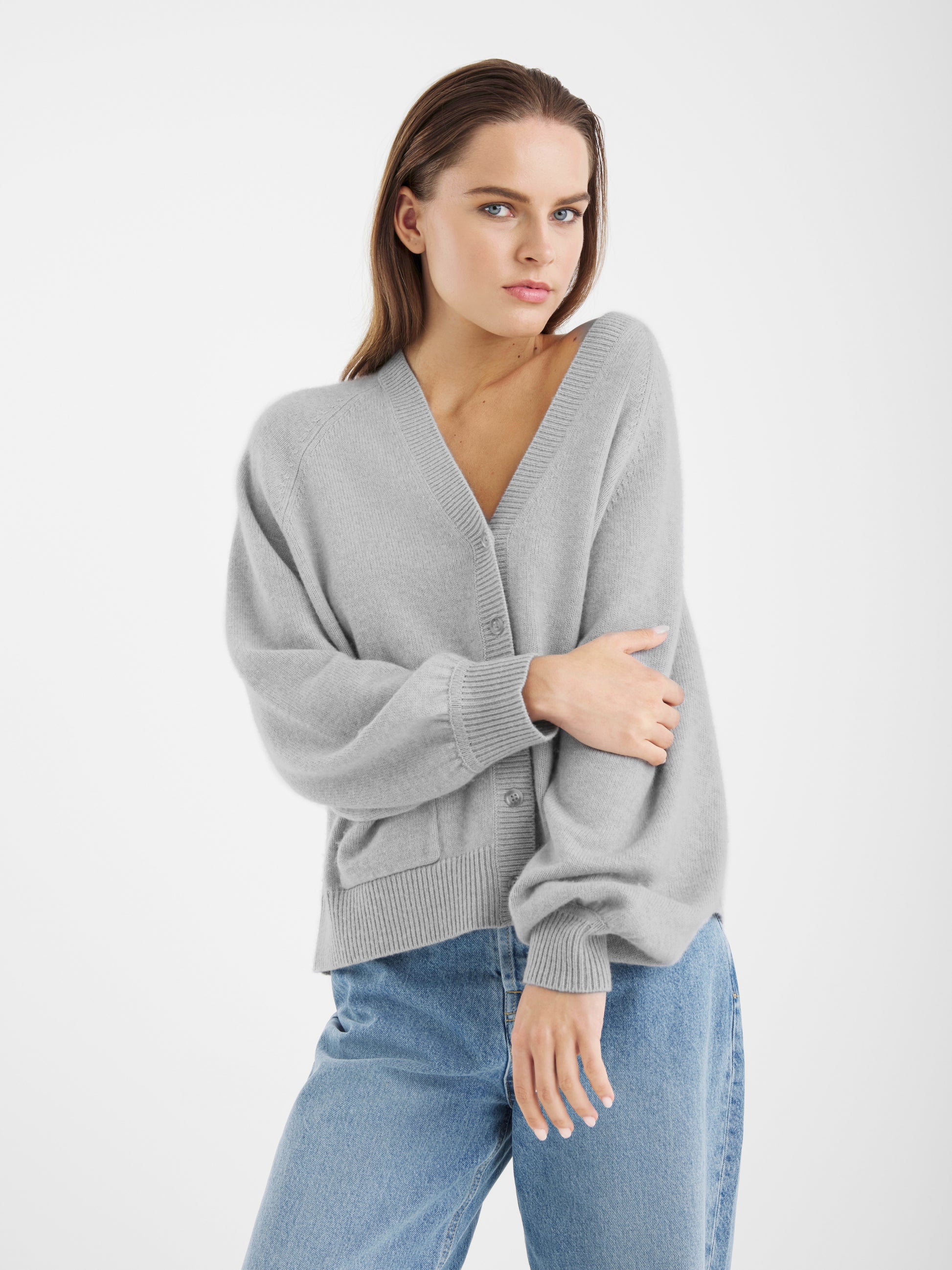 Ein Damen Modell trägt den Bella Cardigan Light Grey und blaue Jeans, steht vor weißem Hintergrund, blickt in die Kamera, hat einen Arm verschränkt und lässt den anderen an ihrer Seite.