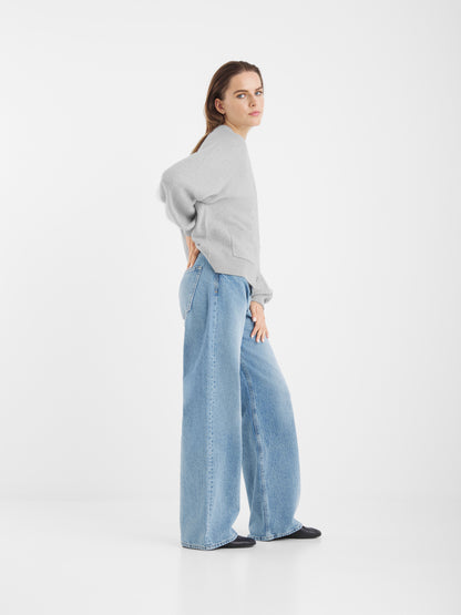 Ein Damen-Model steht seitlich vor einem weißen Hintergrund. Sie trägt die Bella Strickjacke Light Grey, blaue Jeans mit weitem Bein und schwarze Schuhe. Sie hat langes braunes Haar und einen neutralen Gesichtsausdruck, eine Hand ruht auf ihrem unteren Rücken.