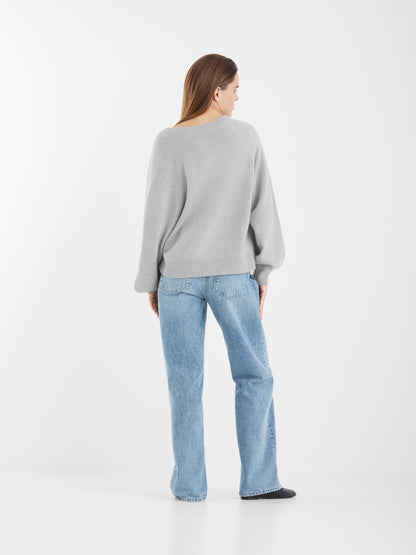 Eine Person mit glattem, hellbraunem Haar blickt von sich weg. Sie trägt den Bella Cardigan Light Grey, hellblaue Jeans und schwarze Schuhe vor einem schlichten weißen Hintergrund.