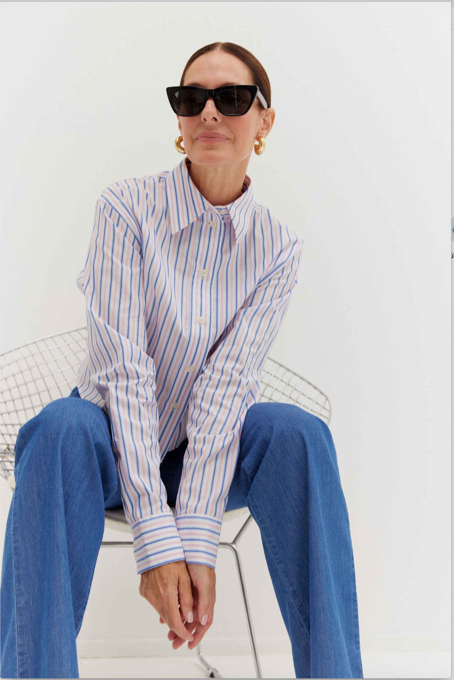 Eine Frau mit schwarzer Sonnenbrille, der Bluse Melbourne Streifen Rose Blau und weit geschnittenen Blue Jeans sitzt auf einem Drahtstuhl vor einem schlichten weißen Hintergrund. Ihr Haar ist hochgesteckt und sie trägt große goldene Ohrringe.