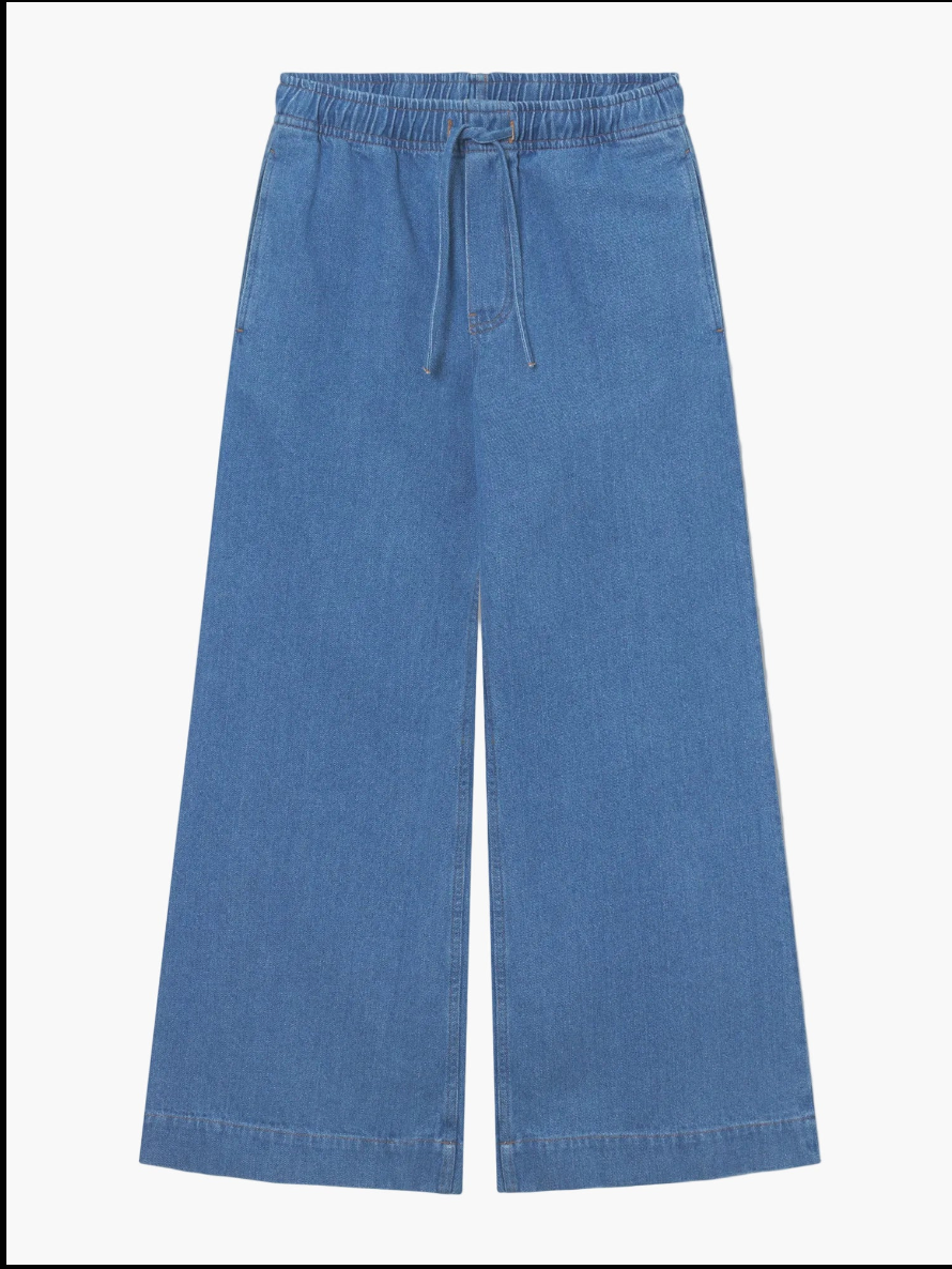 Die Anna Pants Denim sind blaue Jeanshosen mit weitem Bein, elastischem Bund, Kordelzug und Taschen auf der Vorderseite, die flach auf weißem Hintergrund abgebildet sind.