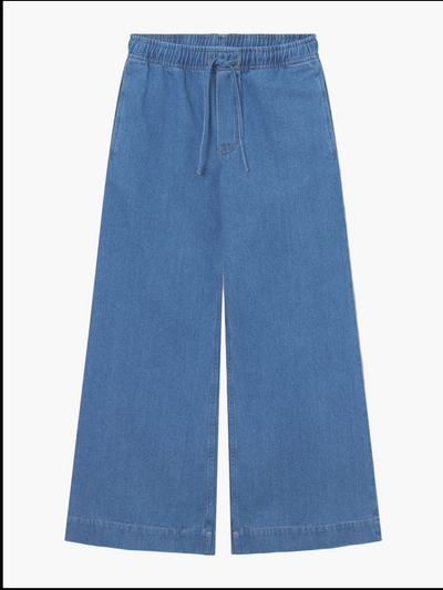 Die Anna Pants Denim sind blaue Jeanshosen mit weitem Bein, elastischem Bund, Kordelzug und Taschen auf der Vorderseite, die flach auf weißem Hintergrund abgebildet sind.