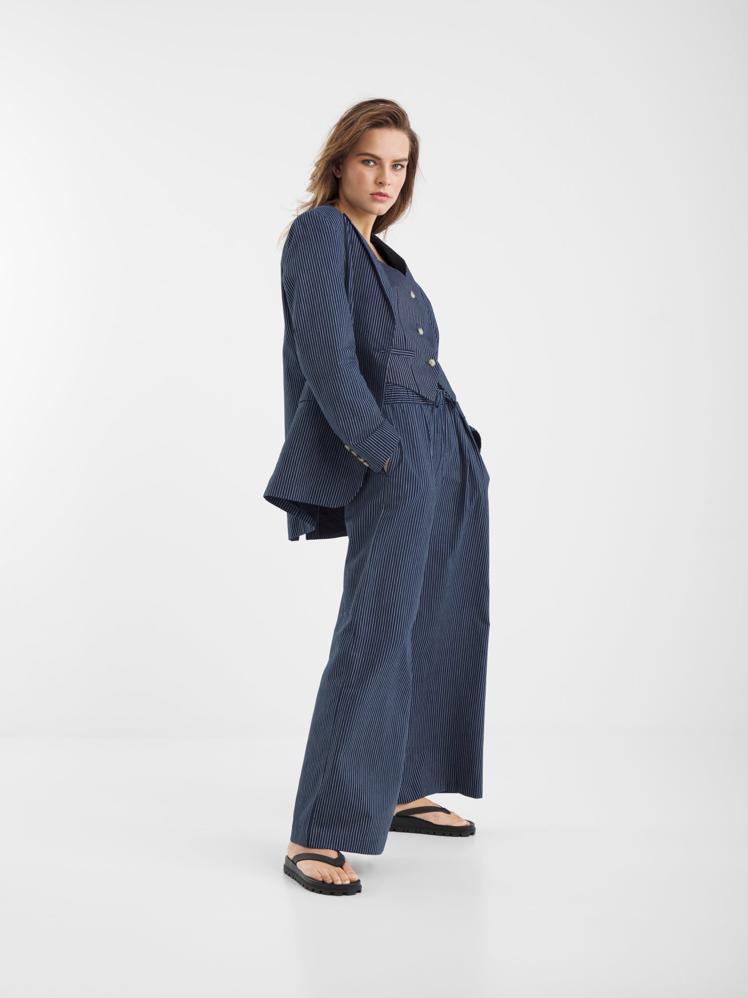 Eine Person steht selbstbewusst in der Pants Pinstripe Navy - einer locker sitzenden Hose mit weitem Bein in Nadelstreifen -, gepaart mit einem doppelreihigen Blazer und schwarzen Sandalen, vor einem schlichten weißen Hintergrund.