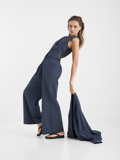 Eine Frau posiert in einem ärmellosen blauen Nadelstreifen-Jumpsuit mit weit geschnittenen Hosen und schwarzen Sandalen und hält den Blazer Pinstripe Navy an ihrer Seite vor einem schlichten weißen Hintergrund.