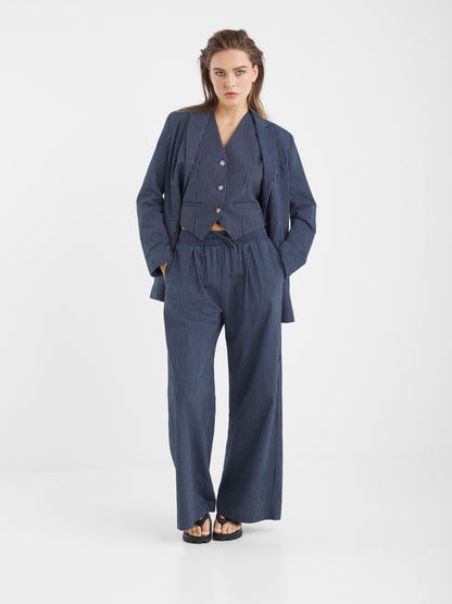 Eine Frau mit langen, gewellten Haaren steht vor einem weißen Hintergrund und trägt den Anzug Vest Pinstripe Navy mit einer Hose mit weitem Bein, einer geknöpften Jacke und schwarzen Sandalen.