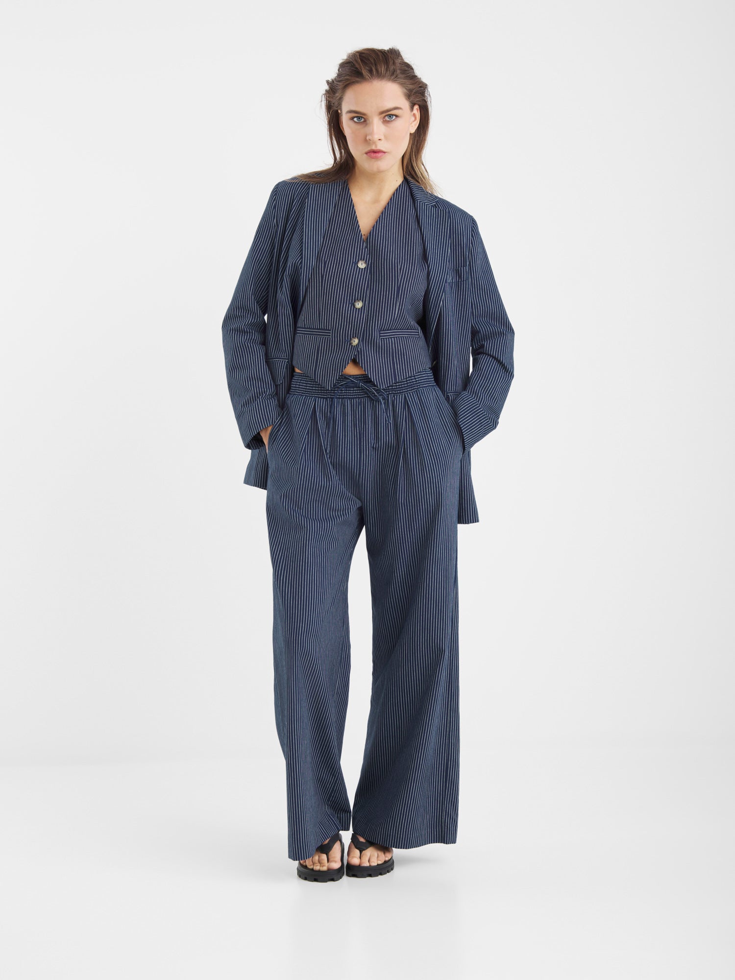 Eine Frau steht vor einem schlichten weißen Hintergrund und trägt den Blazer Pinstripe Navy mit einer Hose mit weitem Bein, eine passende Jacke und schwarze Sandalen. Ihre Hände stecken in den Taschen und ihr Haar ist locker frisiert.