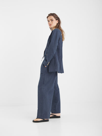 Eine Frau steht seitlich vor einem schlichten weißen Hintergrund und trägt eine Hose mit Nadelstreifen in Marineblau, die mit einer Hose mit weitem Bein und schwarzen Sandalen kombiniert ist.