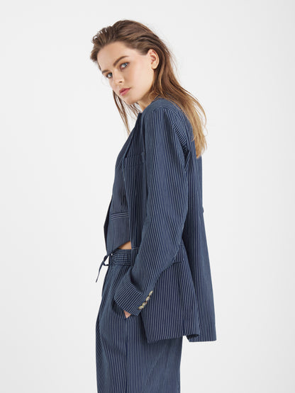 Eine Person mit langem, hellbraunem Haar trägt den Blazer Pinstripe Navy und blickt über die Schulter auf einen schlichten weißen Hintergrund.