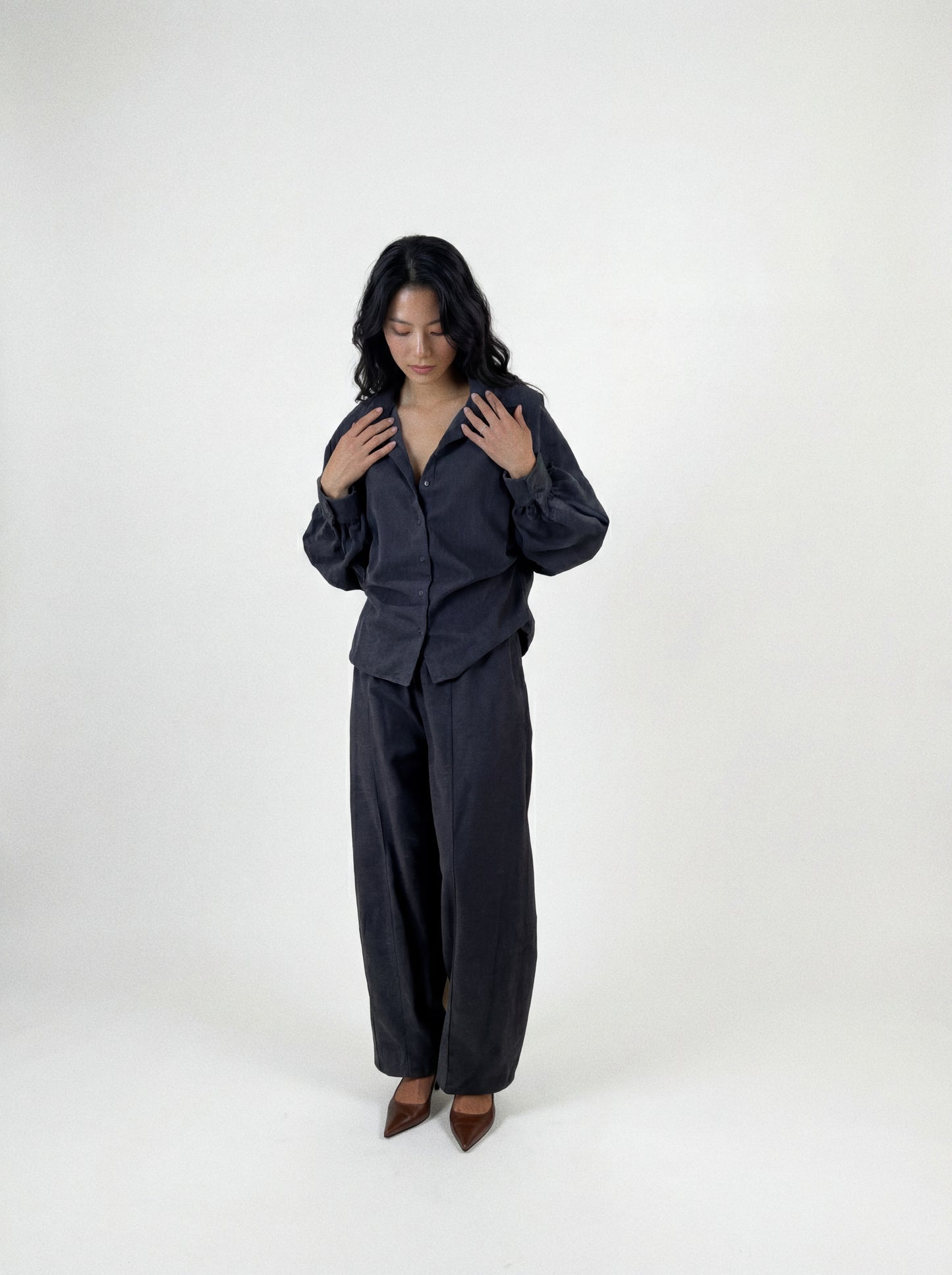 Eine Person mit langen, dunklen Haaren steht vor einem schlichten Hintergrund und trägt eine locker sitzende Bluse „Antonia Slim Flannel Taupe“ sowie eine dazu passende Hose mit weitem Bein aus hochwertiger Baumwolle, dazu braune spitze Schuhe. Sie blickt nach unten, die Hände liegen nahe am Kragen.