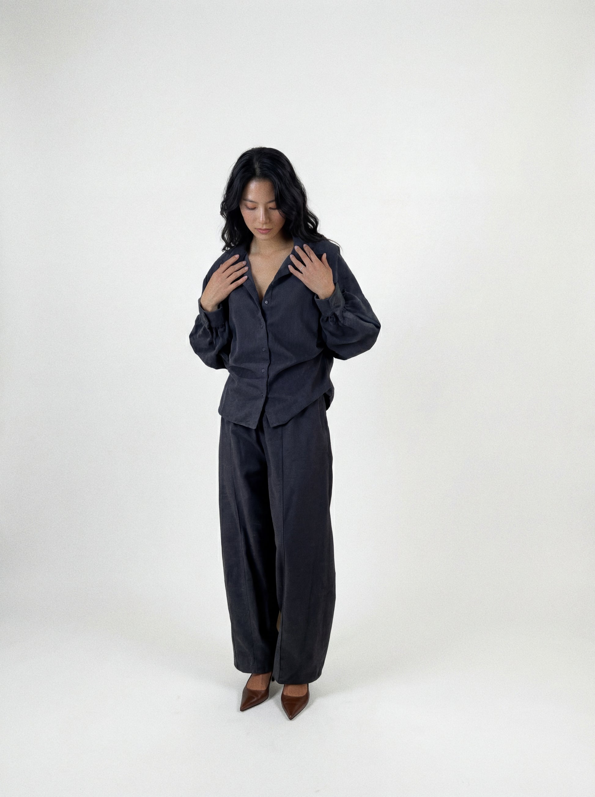 Eine Person mit langen, dunklen Haaren steht vor einem schlichten Hintergrund und trägt eine locker sitzende Bluse „Antonia Slim Flannel Taupe“ sowie eine dazu passende Hose mit weitem Bein aus hochwertiger Baumwolle, dazu braune spitze Schuhe. Sie blickt nach unten, die Hände liegen nahe am Kragen.