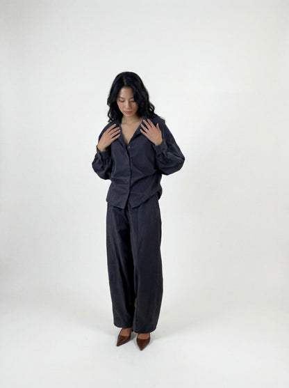 Eine Person mit langen, dunklen Haaren steht vor einem schlichten Hintergrund und trägt eine locker sitzende Bluse „Antonia Slim Flannel Taupe“ sowie eine dazu passende Hose mit weitem Bein aus hochwertiger Baumwolle, dazu braune spitze Schuhe. Sie blickt nach unten, die Hände liegen nahe am Kragen.