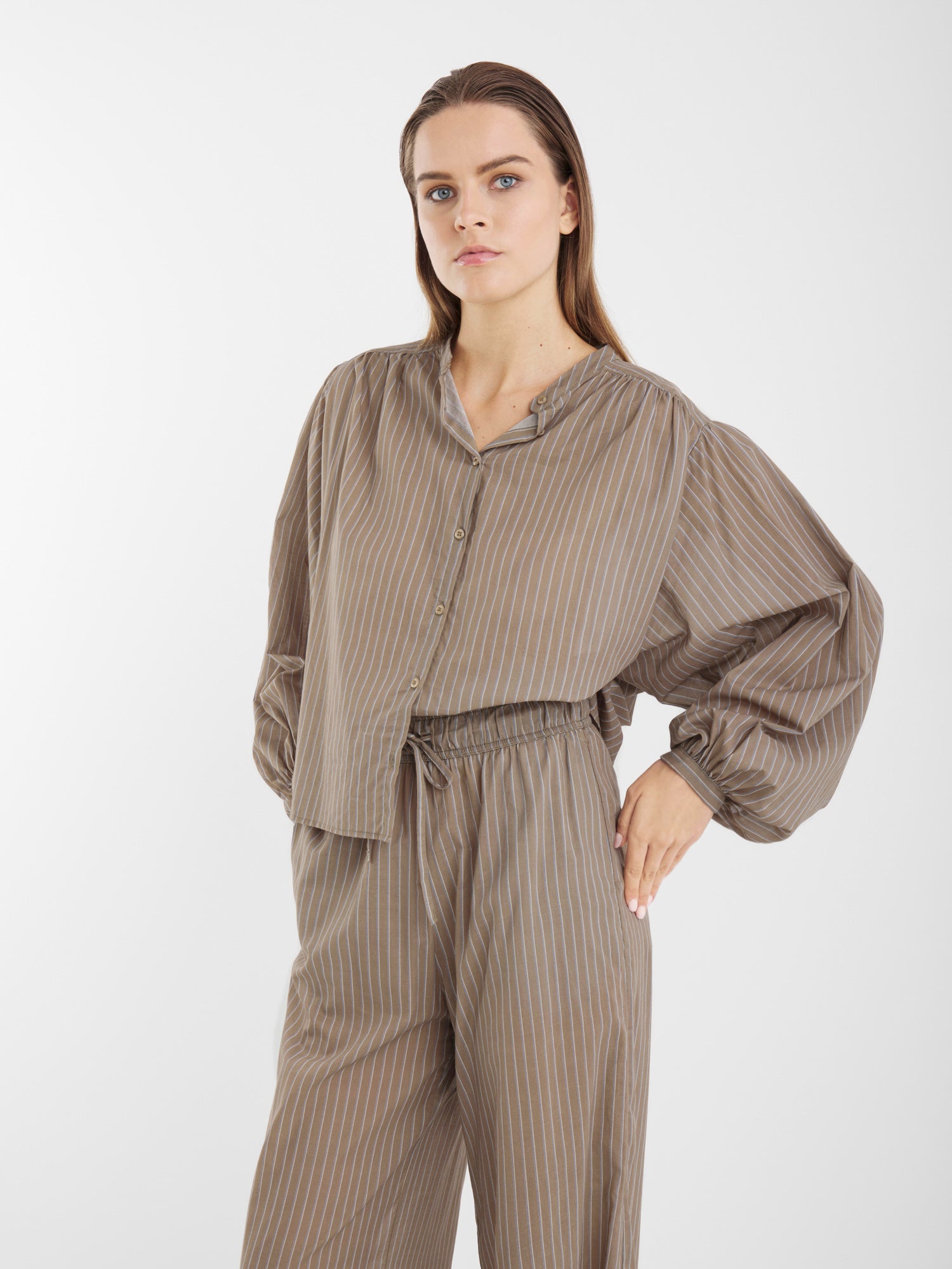 Eine Frau in einer locker sitzenden, langärmeligen Bluse Bella Taupe Stripes und einer dazu passenden Hose mit hoher Taille steht mit einer Hand in der Hüfte vor einem schlichten, hellen Hintergrund.