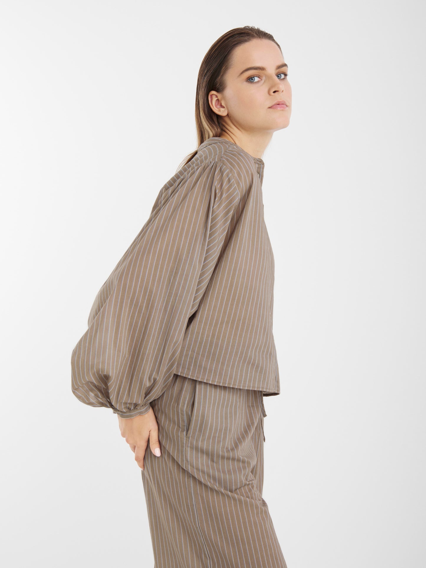 Eine Frau mit glattem, hellbraunem Haar modelliert die Hose Josy Voile Taupe Stripes, gepaart mit einem passenden Oberteil mit Nadelstreifen. Sie steht seitlich und blickt leicht nach oben vor einem schlichten weißen Hintergrund.