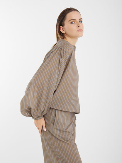 Eine Frau mit glattem, hellbraunem Haar modelliert die Hose Josy Voile Taupe Stripes, gepaart mit einem passenden Oberteil mit Nadelstreifen. Sie steht seitlich und blickt leicht nach oben vor einem schlichten weißen Hintergrund.