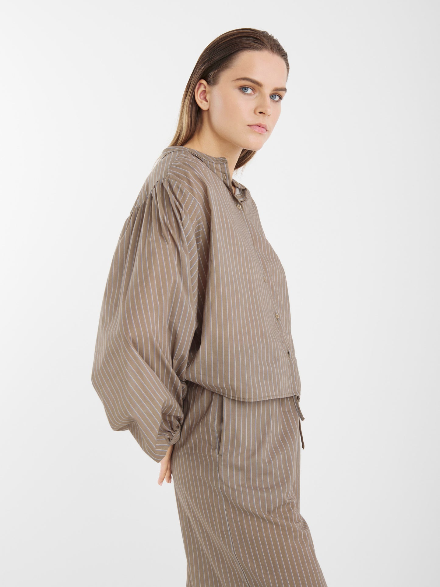 Eine Person mit glattem hellbraunem Haar trägt eine locker sitzende Bluse Bella Taupe Stripes aus Baumwoll-Voile und eine dazu passende Hose mit dünnen weißen Streifen, die vor einem schlichten weißen Hintergrund steht.