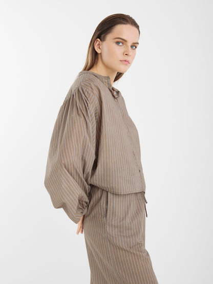 Eine Person mit glattem hellbraunem Haar trägt eine locker sitzende Bluse Bella Taupe Stripes aus Baumwoll-Voile und eine dazu passende Hose mit dünnen weißen Streifen, die vor einem schlichten weißen Hintergrund steht.