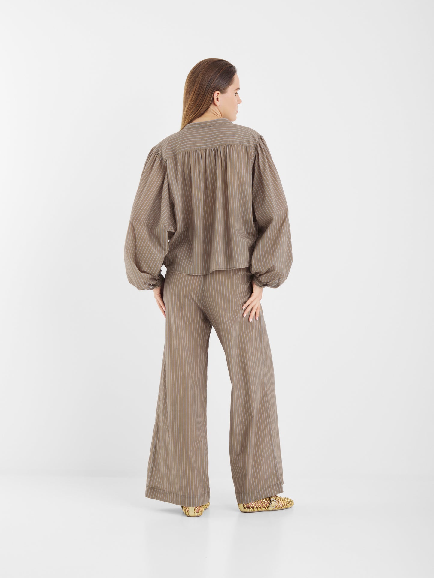 Eine Frau mit langen Haaren steht mit dem Pants Josy Voile Taupe Stripes - einem locker sitzenden, taupefarbenen Nadelstreifen-Hosenanzug mit weitem Bein, einer Bluse mit voluminösen Ärmeln und beigefarbenen, gemusterten Schuhen - vor einem schlichten weißen Hintergrund.
