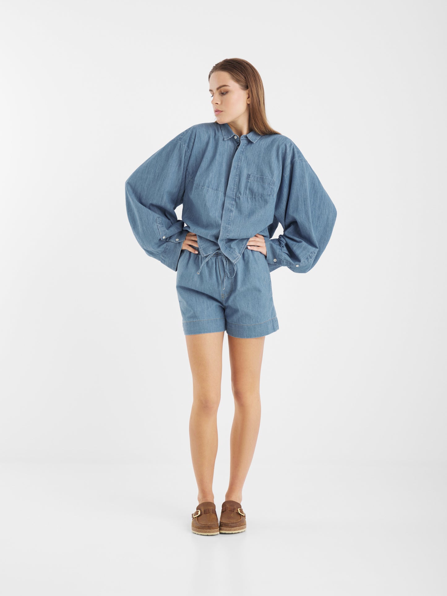 Eine Frau steht in einem Studio mit weißem Hintergrund, trägt ein locker sitzendes, langärmeliges blaues Hemd und eine kurze dunkle Jeans und schaut mit den Händen in den Hüften zur Seite.