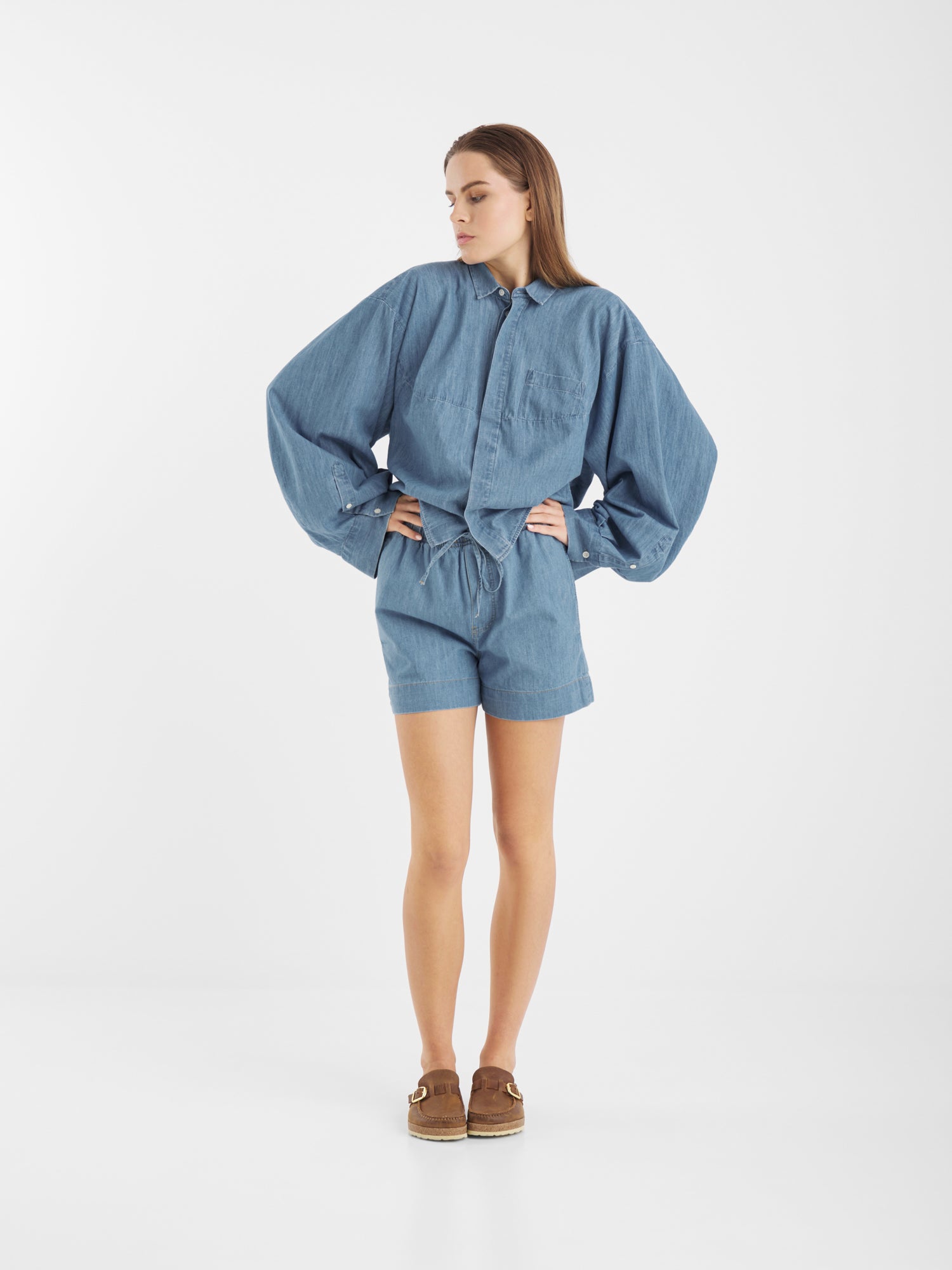 Eine Frau steht in einem Studio mit weißem Hintergrund, trägt ein locker sitzendes, langärmeliges blaues Hemd und eine kurze dunkle Jeans und schaut mit den Händen in den Hüften zur Seite.