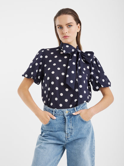 Eine Frau steht mit den Händen in den Jeanstaschen und trägt die Bluse Bow Slim Dots Navy Off White mit weißen Tupfen, kurzen Puffärmeln und einer großen Schleife im Nacken. Ihr glattes Haar hat sie hinter die Ohren gesteckt.