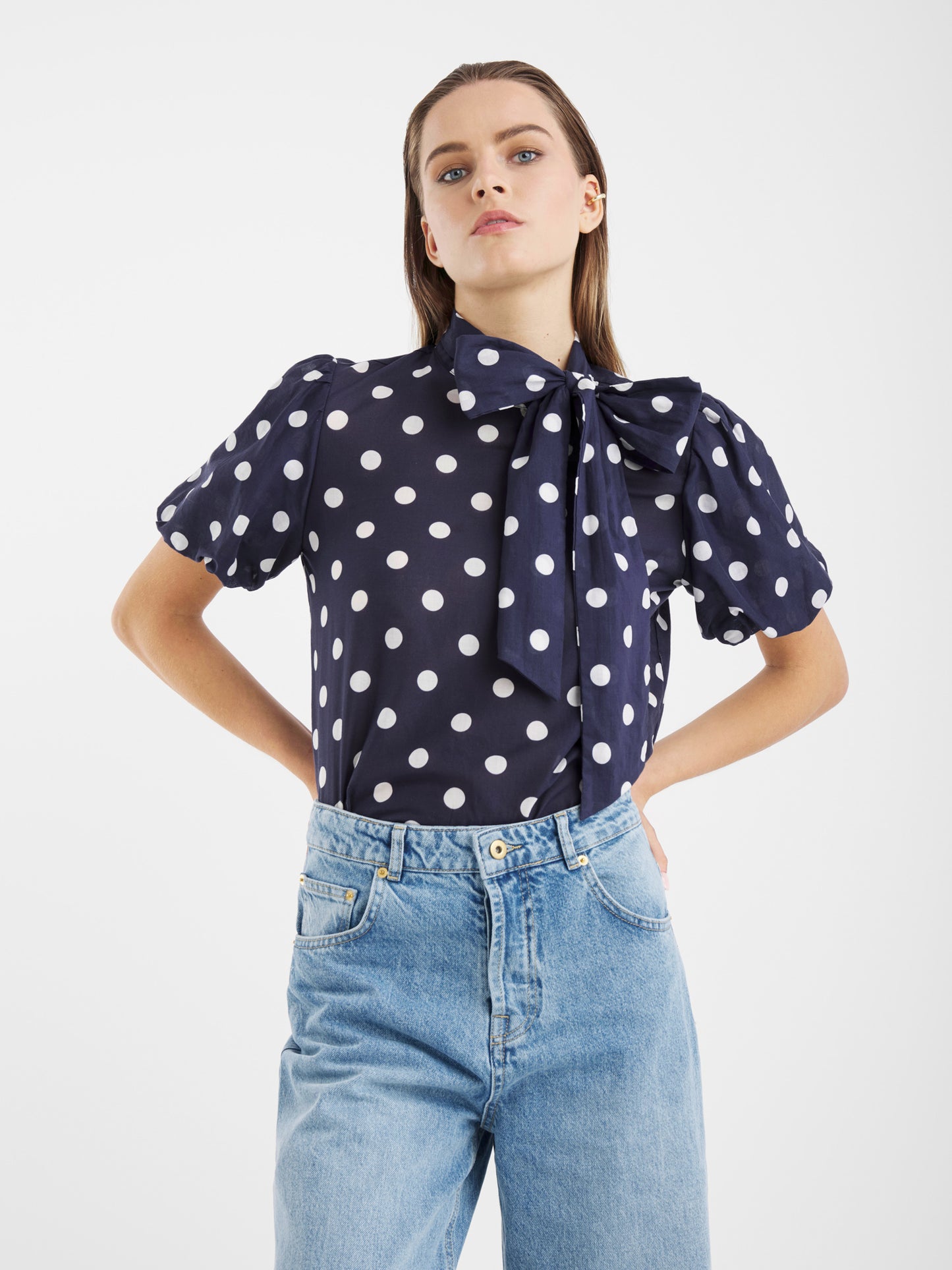 Eine Frau steht vor einem einfarbig weißen Hintergrund und trägt die Bluse Bow Slim Dots Navy Off White mit kurzen Puffärmeln, weißen Tupfen und einer großen Schleife im Nacken, gepaart mit hellblauen High-Waisted Jeans.