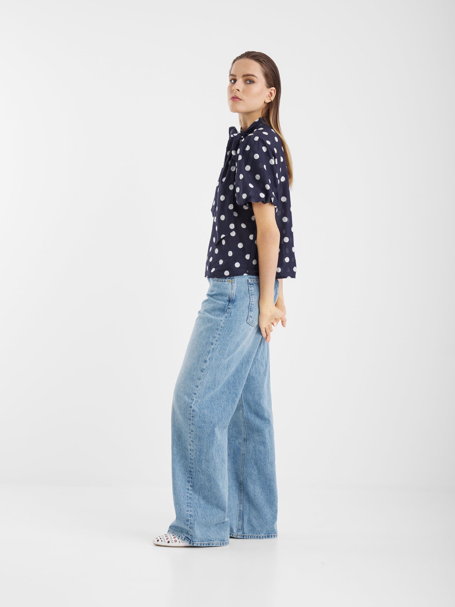 Eine Frau im Profil trägt die Bluse Bow Slim Dots Navy Off White mit hellblauen, weit geschnittenen Jeans. Sie hat glattes braunes Haar, einen neutralen Ausdruck und steht vor einem schlichten weißen Hintergrund.