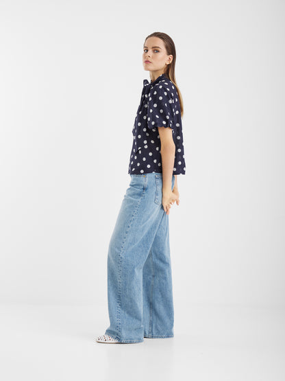 Eine Frau im Profil trägt die Bluse Bow Slim Dots Navy Off White mit hellblauen, weit geschnittenen Jeans. Sie hat glattes braunes Haar, einen neutralen Ausdruck und steht vor einem schlichten weißen Hintergrund.