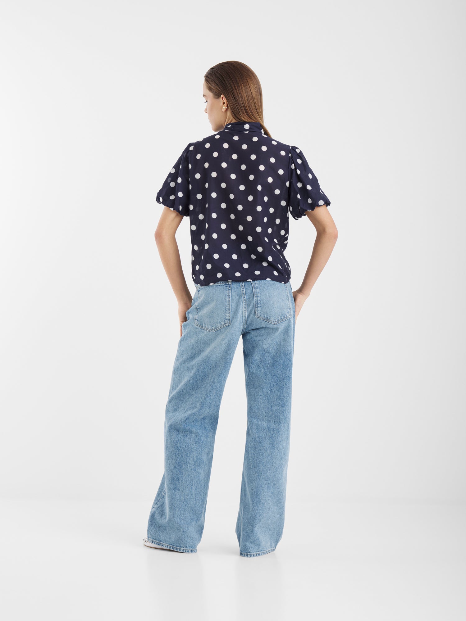 Eine Frau steht mit der Bluse Bow Slim Dots Navy Off White und hellblauen, weit geschnittenen Jeans vor einem schlichten weißen Hintergrund.