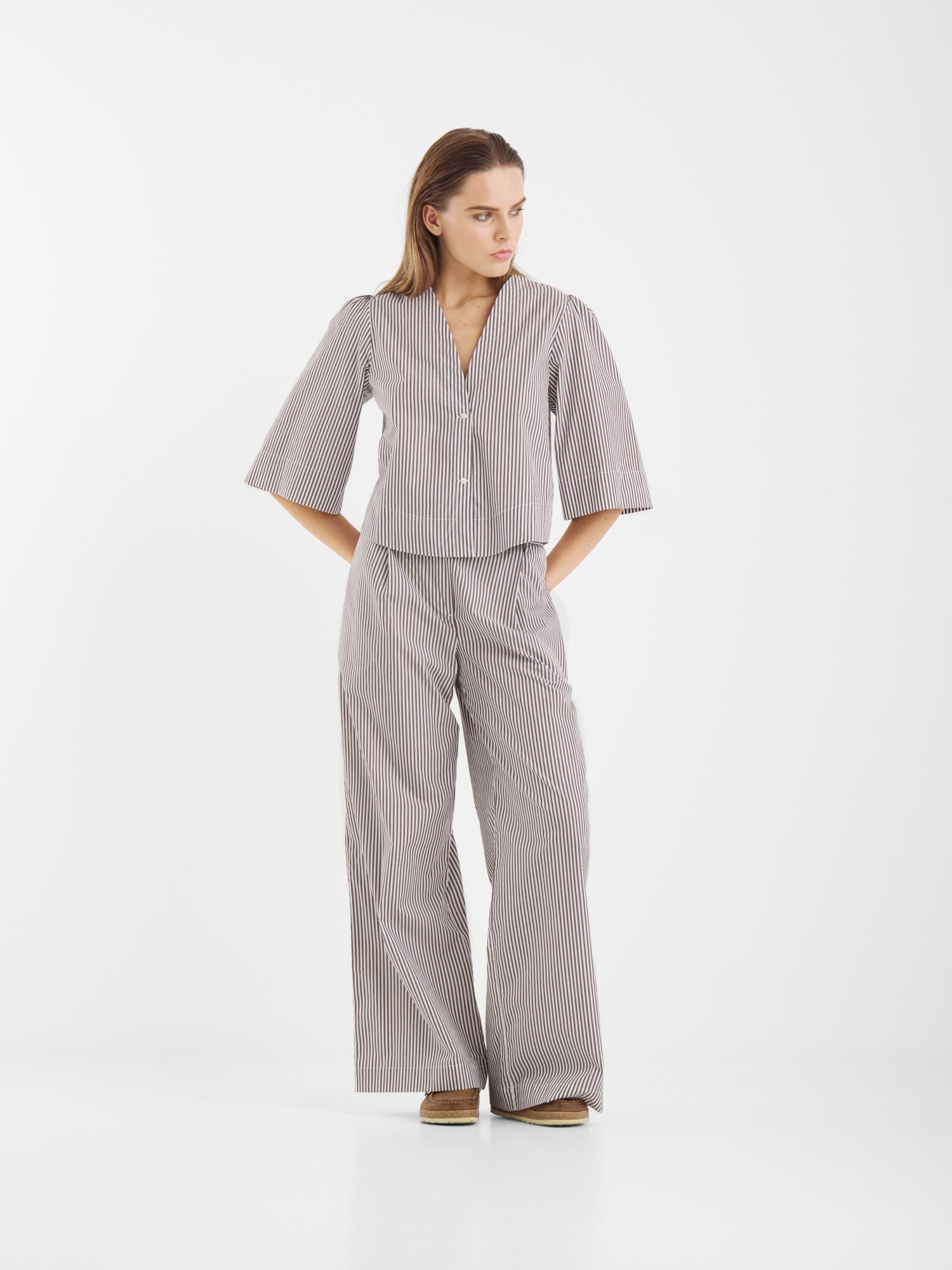 Eine Frau steht vor einem weißen Hintergrund und trägt die Bluse California Brown Stripes mit einer weit geschnittenen Hose. Mit den Händen in den Hosentaschen blickt sie zur Seite und unterstreicht damit die entspannte, stilvolle Haltung.