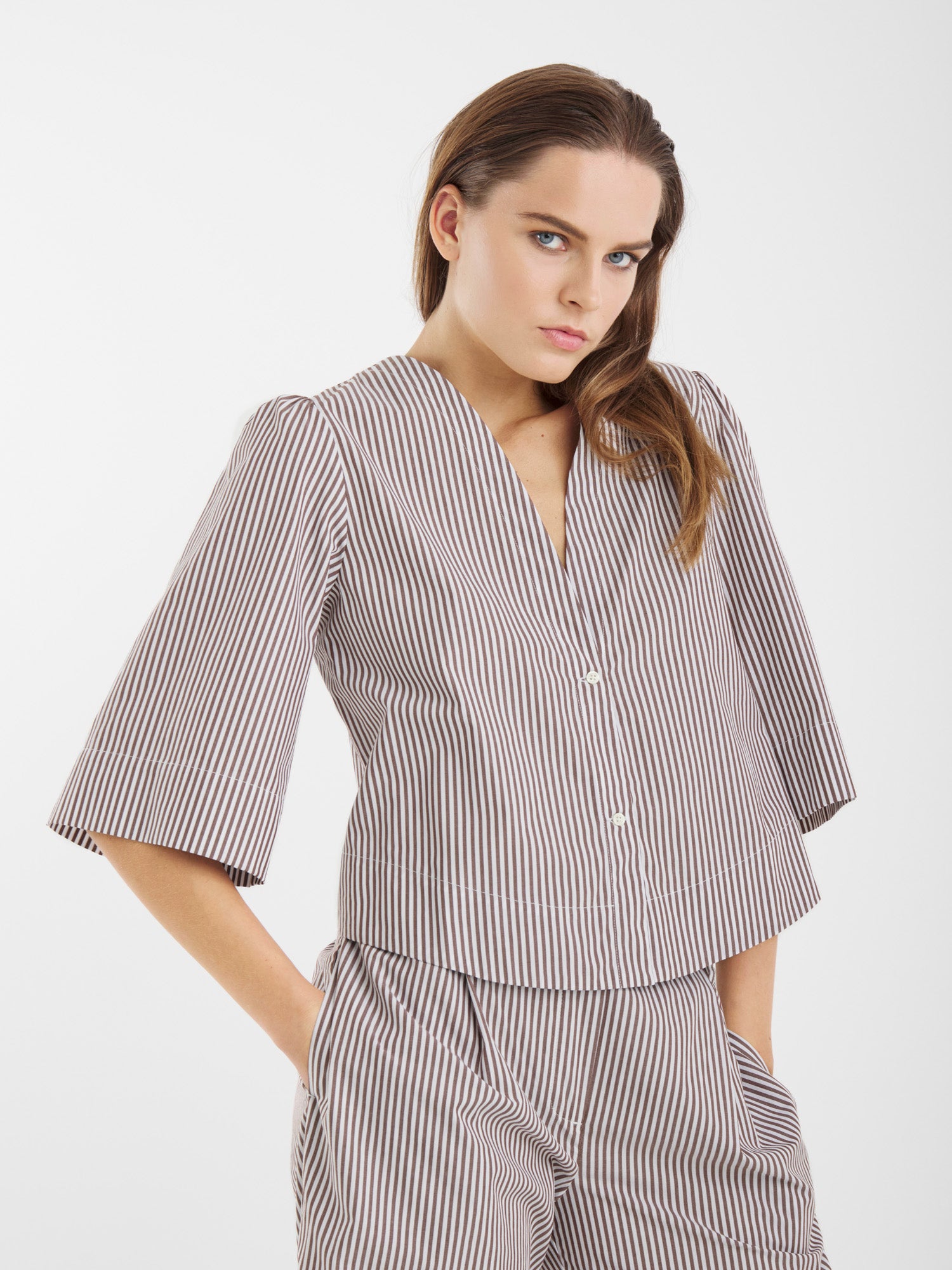 Eine Frau trägt die Bluse California Brown Stripes mit ellbogenlangen Ärmeln und einer dazu passenden Hose mit hoher Taille. Sie steht mit den Händen in den Taschen und blickt selbstbewusst in die Kamera vor einem schlichten weißen Hintergrund.