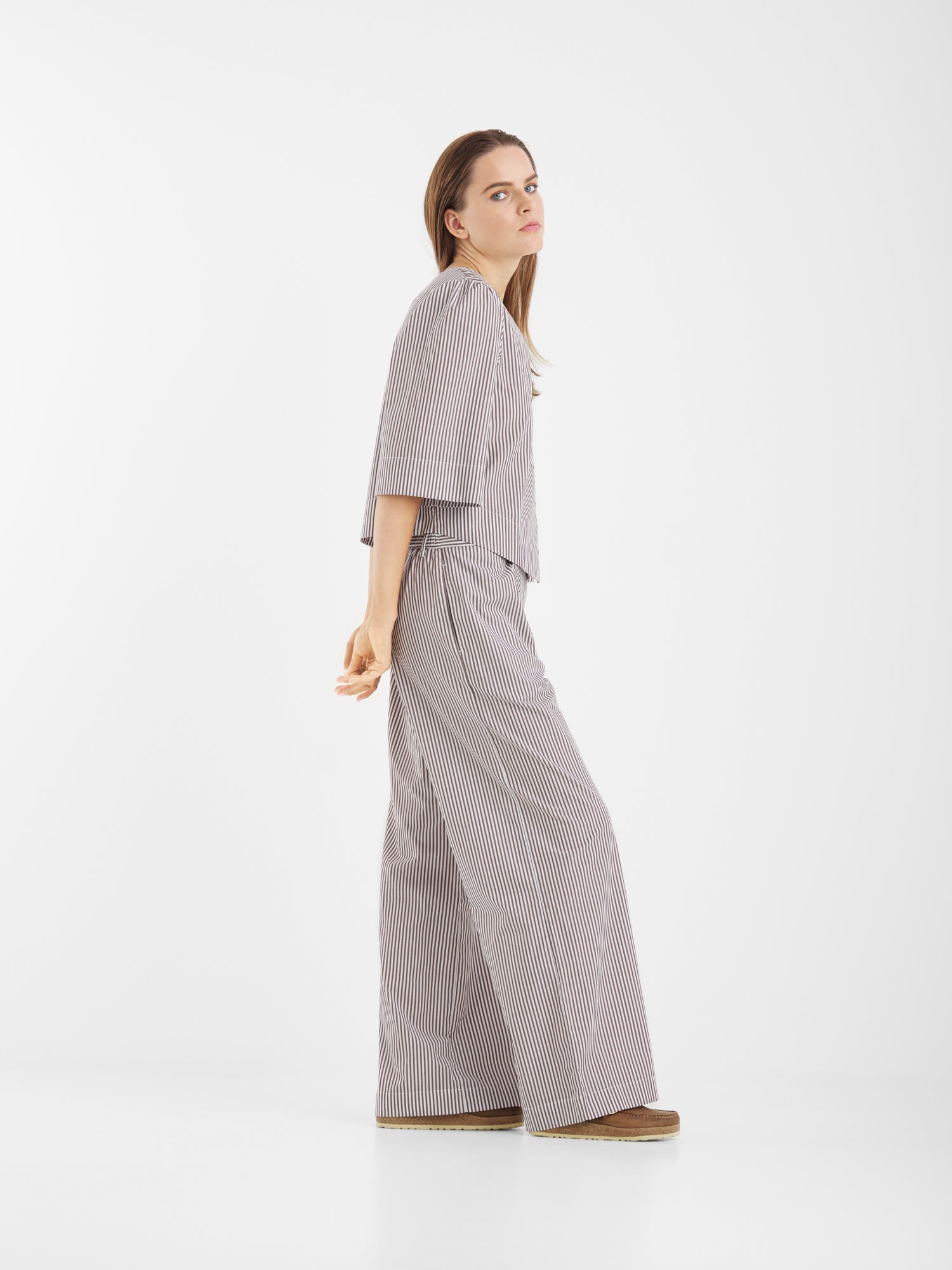 Eine Frau steht seitwärts in einer minimalistischen, weißen Umgebung und trägt die Bluse California Brown Stripes mit einer Hose mit weitem Bein und Plateauschuhen. Ihr langes Haar und ihr neutraler Gesichtsausdruck ergänzen die klare Kulisse.