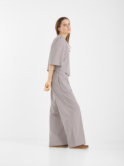 Eine Frau steht seitwärts in einer minimalistischen, weißen Umgebung und trägt die Bluse California Brown Stripes mit einer Hose mit weitem Bein und Plateauschuhen. Ihr langes Haar und ihr neutraler Gesichtsausdruck ergänzen die klare Kulisse.