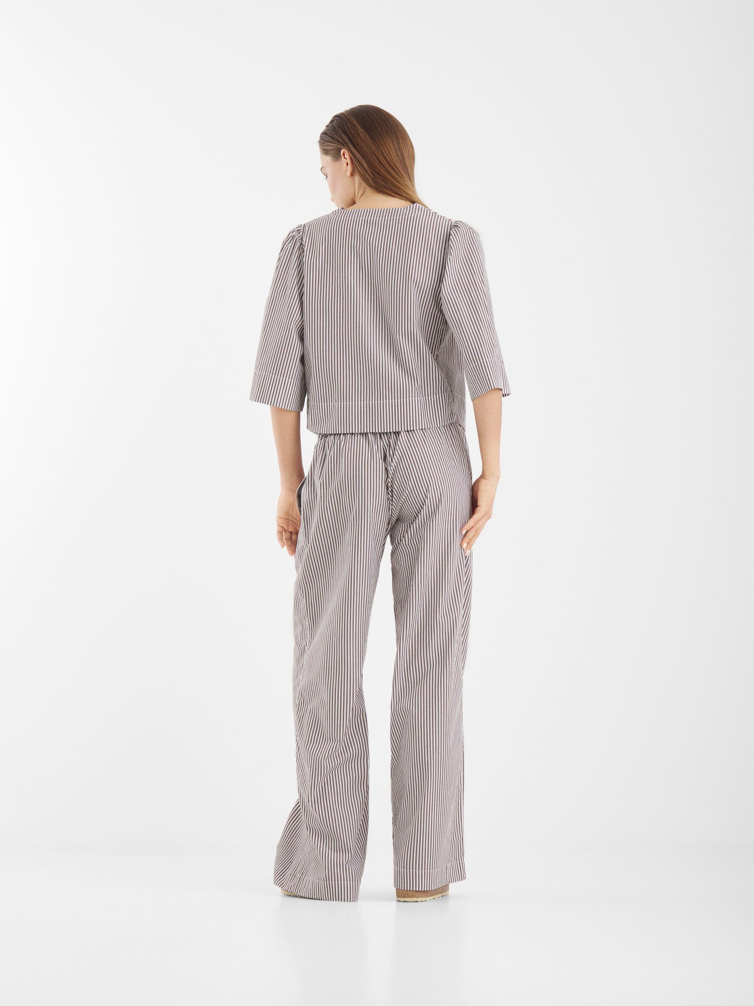 Eine Frau steht mit dem Gesicht zur Wand und trägt die Bluse California Brown Stripes mit einer weit geschnittenen Hose. Das langärmelige, locker sitzende Oberteil ist mit braunen Streifen versehen und steht vor einem schlichten weißen Hintergrund.