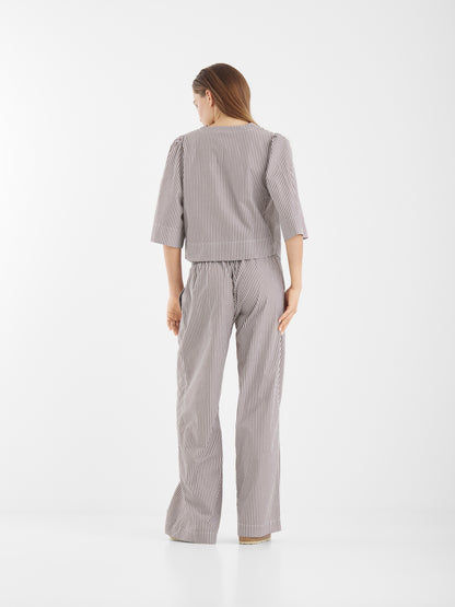 Eine Frau steht mit dem Gesicht zur Wand und trägt die Bluse California Brown Stripes mit einer weit geschnittenen Hose. Das langärmelige, locker sitzende Oberteil ist mit braunen Streifen versehen und steht vor einem schlichten weißen Hintergrund.