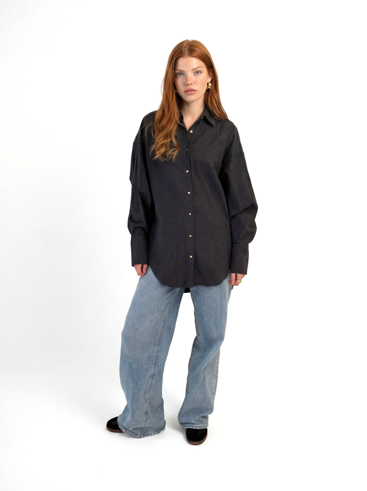 Eine Person steht vor einem weißen Hintergrund und trägt die Bluse „Carla Anthra“ – ein locker sitzendes, dunkles Hemd mit Knöpfen –, dazu hellblaue Jeans mit weitem Bein und schwarze Schuhe. Sie hat langes, rotbraunes Haar und einen neutralen Gesichtsausdruck.