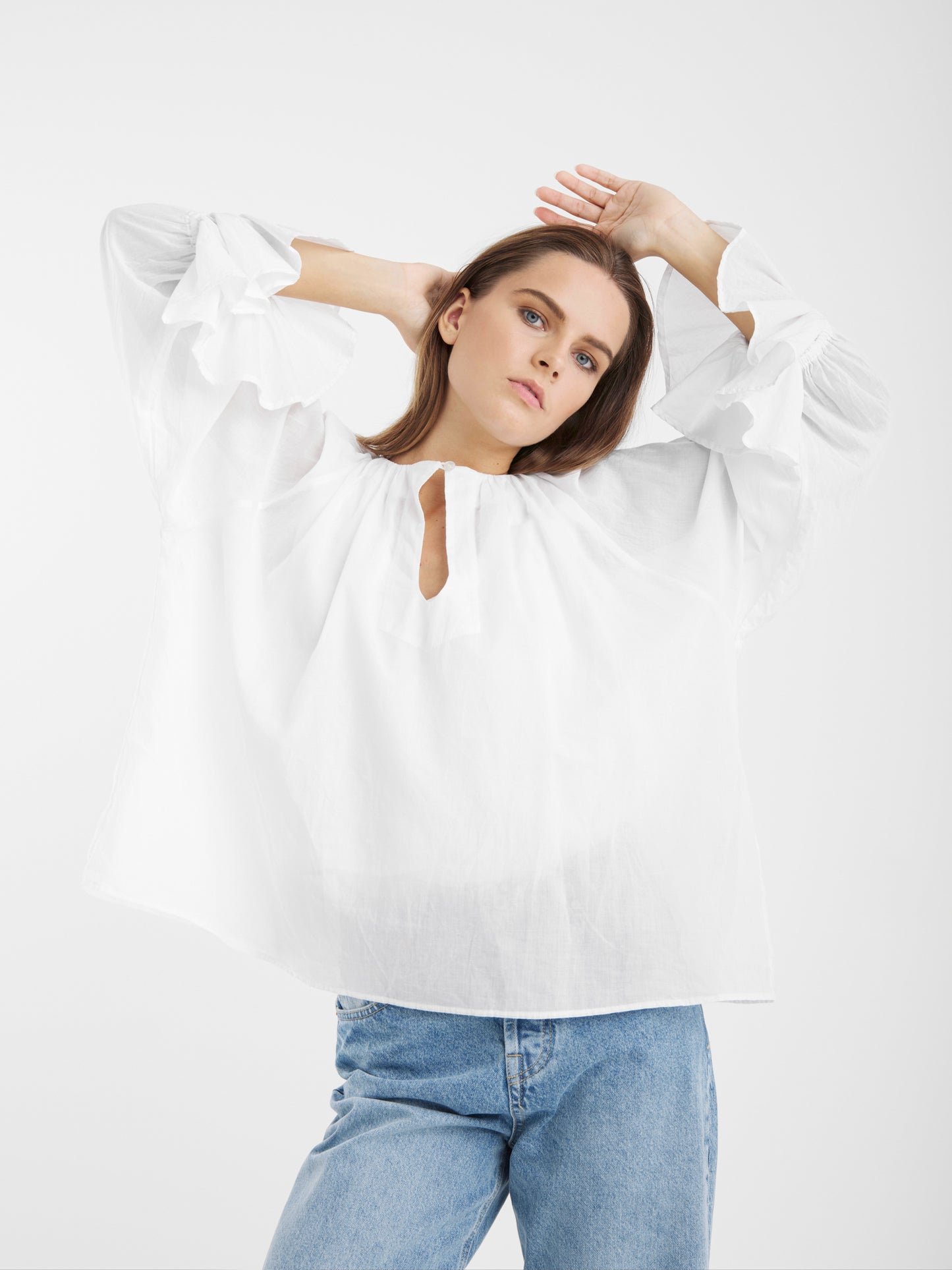 Eine Frau posiert mit über den Kopf erhobenen Armen. Sie trägt die Bluse Carmen White mit gerüschten Ärmeln und hellblaue Jeans vor einem schlichten weißen Hintergrund.