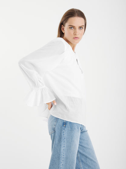 Eine Frau mit glattem braunem Haar steht im Seitenprofil vor einem schlichten weißen Hintergrund und trägt die Bluse Carmen White - eine lockere langärmelige Bluse mit gerüschten Manschetten - und hellblaue Jeans.