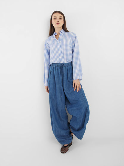 Eine Frau mit langem glattem Haar steht vor einem schlichten weißen Hintergrund. Sie trägt die Bluse Casual Check Blue White Check und eine weite, blaue Hose, gepaart mit schwarzen Netzschuhen.